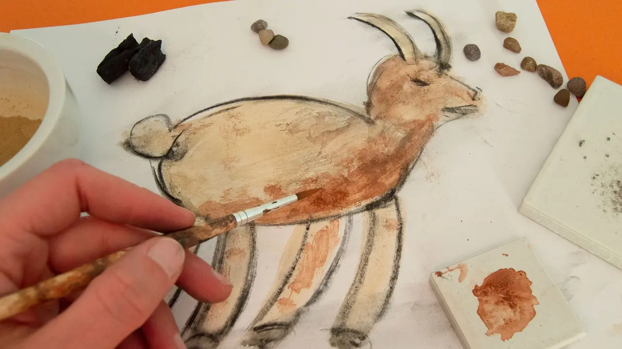 Workshop Malen mit Steinen Eine Hand mit Pinsel malt eine Ziege. Auf dem Papier liegen verschiedene Steine.